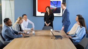 A group of people in a meeting room gathered around a table, with a presentation screen in the background displaying a graphic representation.