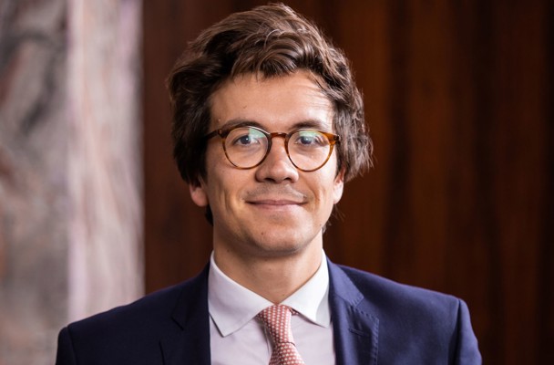 Mathias in a dark blazer and light-colored tie, with a neutral expression, sitting against a warm wooden background.