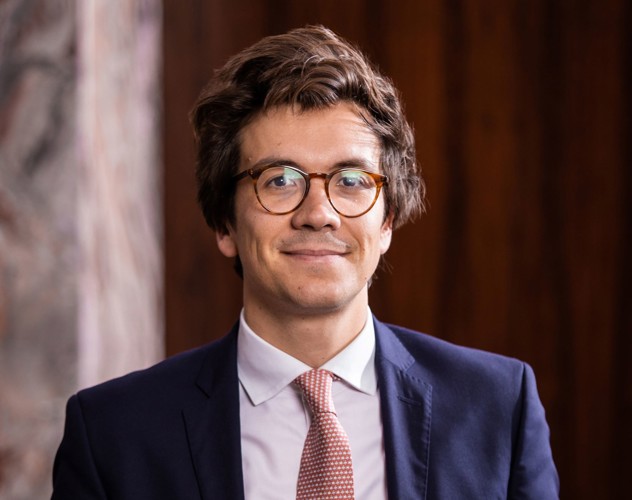 Mathias in a dark blazer and light-colored tie, with a neutral expression, sitting against a warm wooden background.