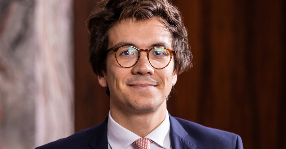 Mathias in a dark blazer and light-colored tie, with a neutral expression, sitting against a warm wooden background.