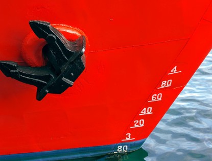 Zicht op een helderrood schip met een zwart anker en dieptemeter waarop diepteniveaus in witte cijfers zijn gemarkeerd.