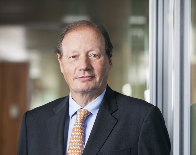 Mr D.M. Sluimers in a black suit with an orange tie, standing in front of a modern office wall, with a blurred background.