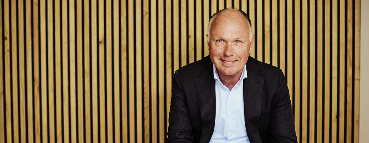 Nick in a black jacket and white shirt is sitting on a bench against a background of vertical wooden slats.