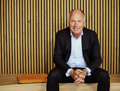 Nick in een zwart jasje en wit hemde zit op een bank voor een achtergrond van verticale houten latten.