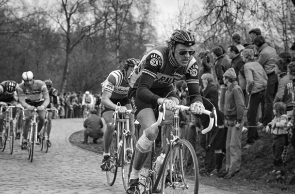 A group of cyclists in colorful jerseys, determined on a cobblestone road, surrounded by an enthusiastic crowd in the background.