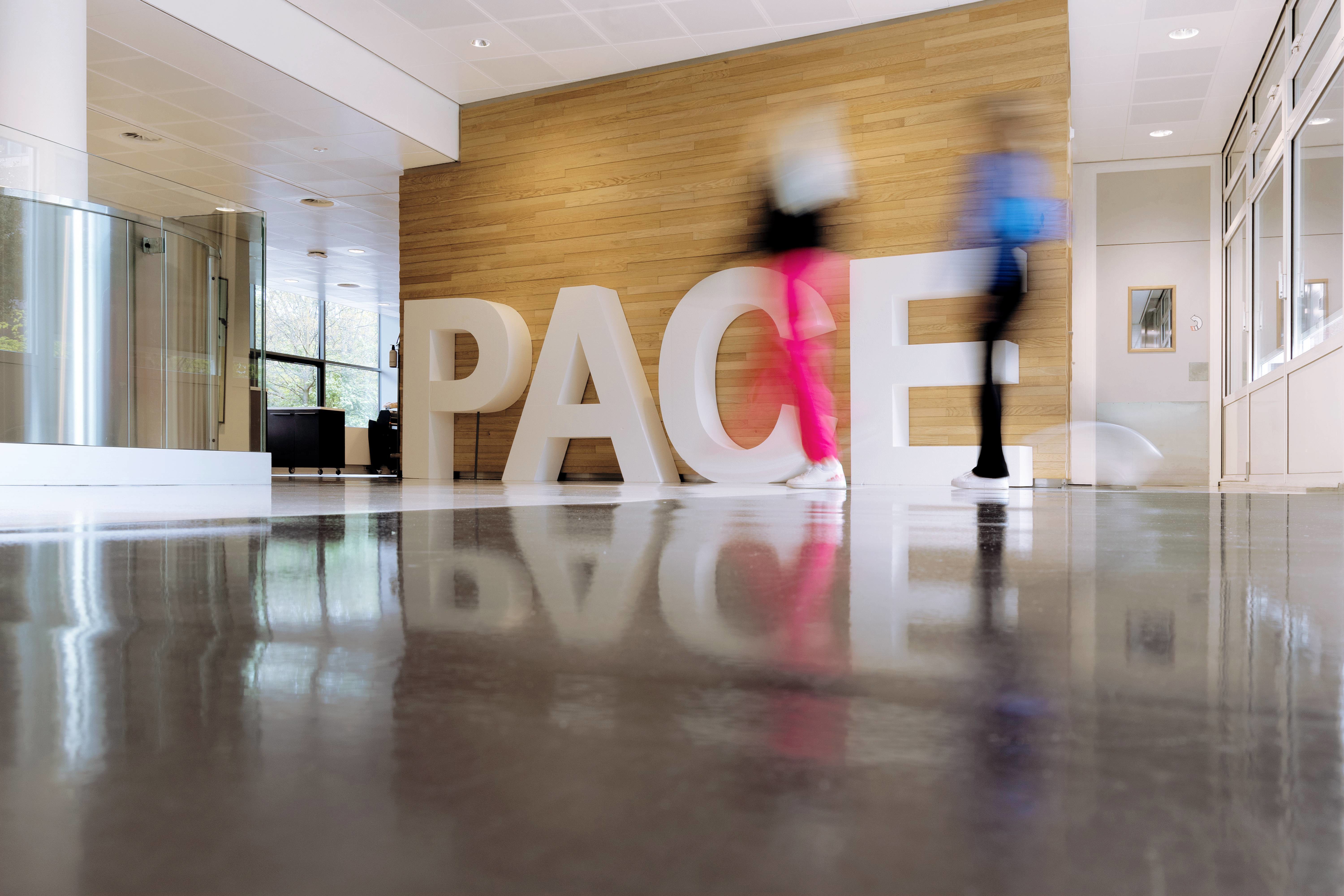 Two people walk past large white letters spelling the word 'PACE' in a modern lobby.