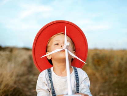 A child in a red hat stands in a field of yellow grass, looking playful with an arrow in its mouth.