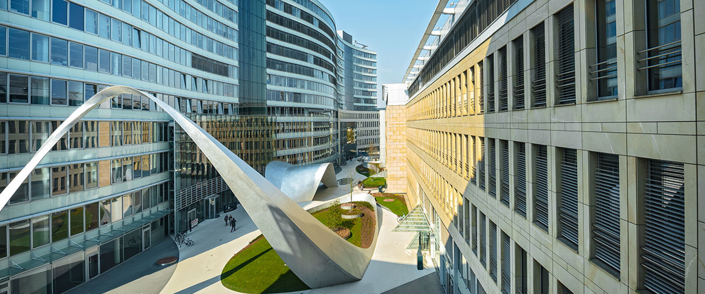 Modern architectural illustration of an open courtyard featuring sculptures and green spaces between buildings with glass and stone facades.