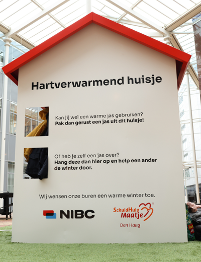 Wit kunstobject in de vorm van een huisje met de tekst Hartverwarmend huisje. Het object maakt deel uit van een oproep voor winterjassen en benadrukt samenwerking. Onderaan staan de logo's van NIBC en SchuldHulpMaatje Den Haag.