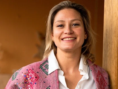 Roxane in a colorful knitted cardigan leans against a wooden pillar, smiling and looking relaxed.