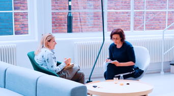 Twee vrouwen zitten in een lichte, moderne ruimte. De ene heeft een notitieboekje en de andere een pen, terwijl ze met elkaar praten.