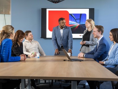 Groep mensen in een vergadering, waarbij een man in het midden een presentatie geeft voor een scherm met grafieken.
