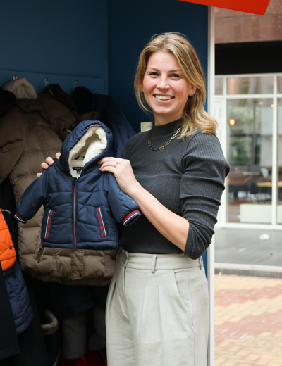Vrouw staat voor een rek met jassen in een winkel en houdt een blauw babyjasje met capuchon vast.