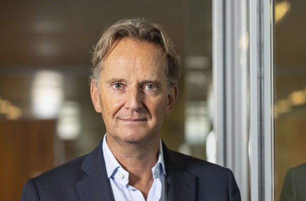 Joop in a blazer stands in front of a glass wall, looking at the camera with a professional demeanor.