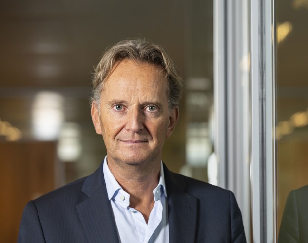 Joop in een blazer staat voor een glazen wand en kijkt naar de camera met een professionele uitstraling.