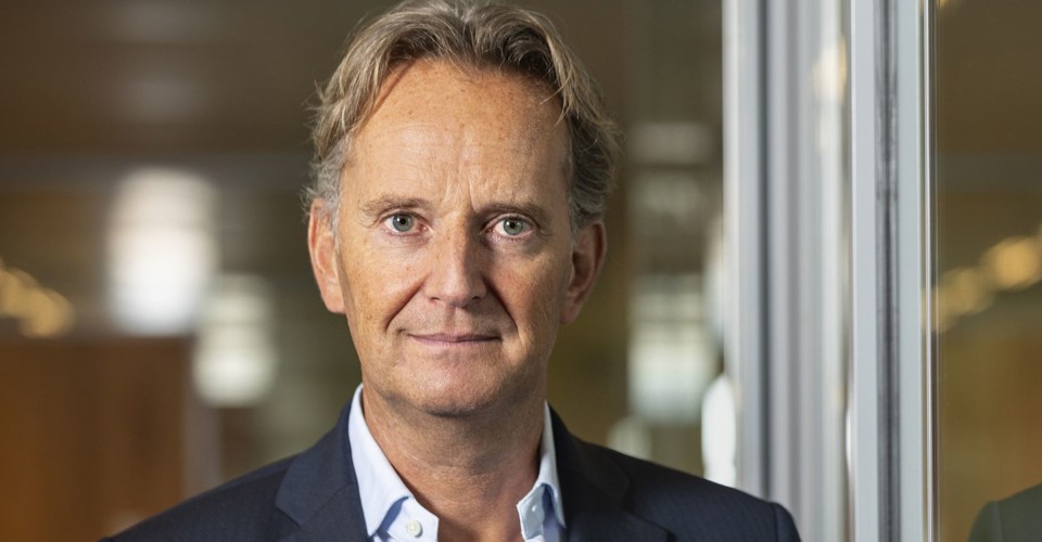 Joop in een blazer staat voor een glazen wand en kijkt naar de camera met een professionele uitstraling.