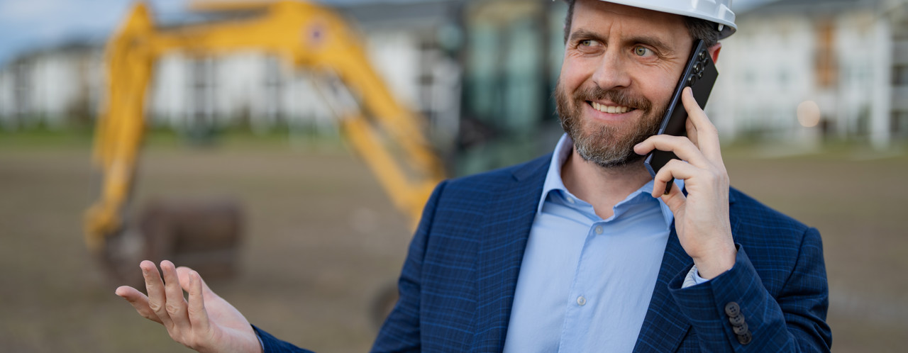 A man in a blazer stands on a construction site, speaking on his phone, with an excavator visible in the background.