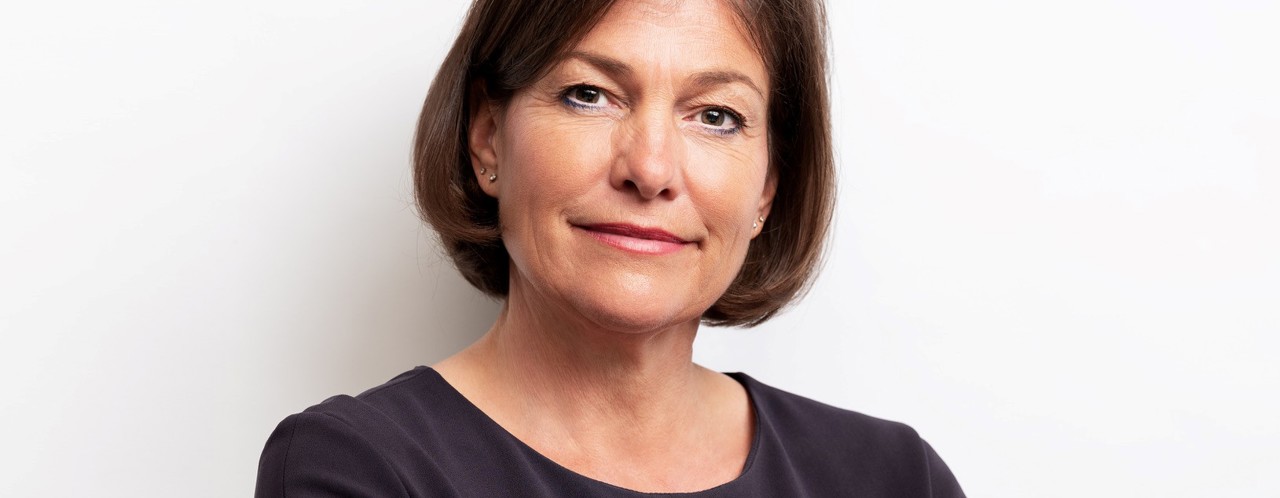 Leni with a neutral expression, wearing a dark blue top, stands with her arms crossed against a light background.