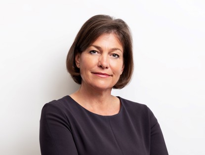 Leni with a neutral expression, wearing a dark blue top, stands with her arms crossed against a light background.