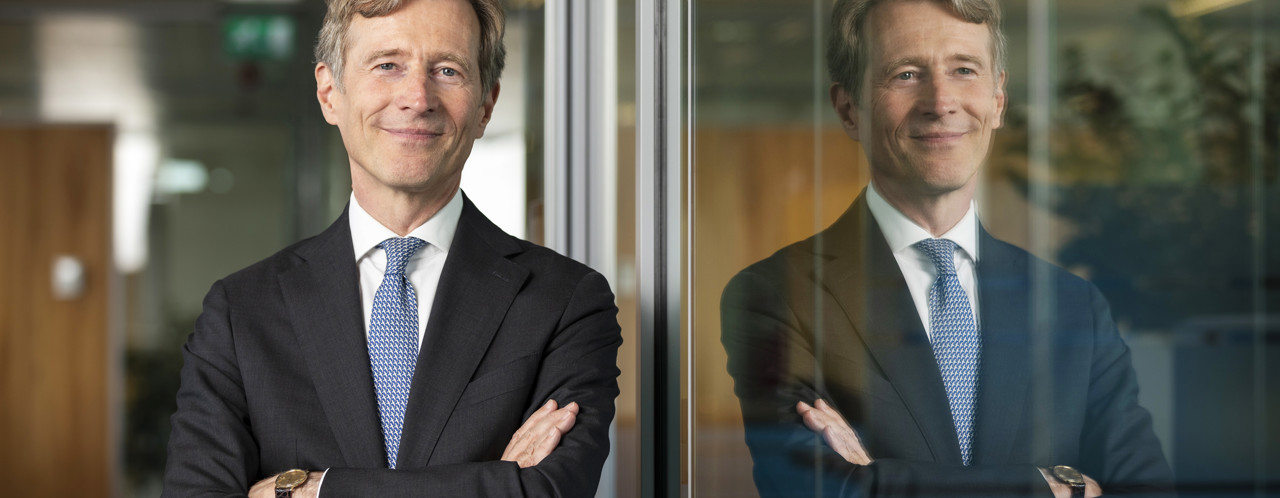 Jeroen in een zwart pak met een blauwe stropdas staat met gekruiste armen voor een glazen wand, die het beeld weerkaatst.