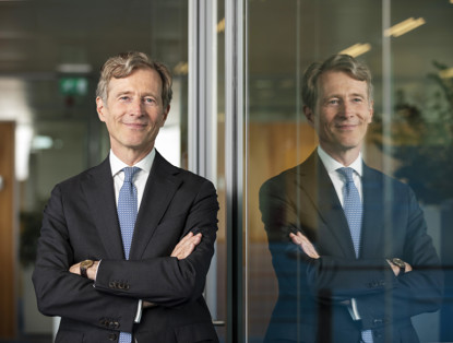 Jeroen in een zwart pak met een blauwe stropdas staat met gekruiste armen voor een glazen wand, die het beeld weerkaatst.