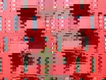 Rood gebouw met grote ramen en twee uitgeronde balkons, omringd door groene planten.