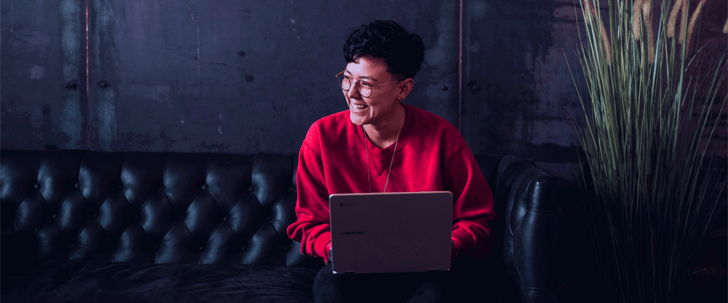 Een persoon in een rode trui zit op een bank met een laptop op schoot, voor een donkere, industriƫle achtergrond.