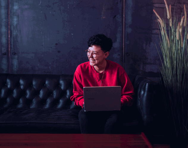 Een persoon in een rode trui zit op een bank met een laptop op schoot, voor een donkere, industriƫle achtergrond.