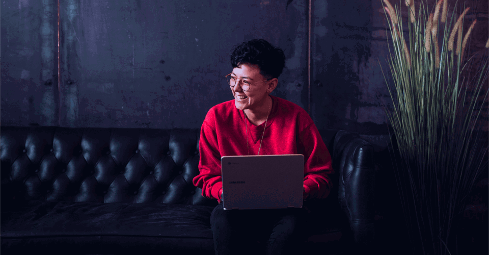 Een persoon in een rode trui zit op een bank met een laptop op schoot, voor een donkere, industriƫle achtergrond.