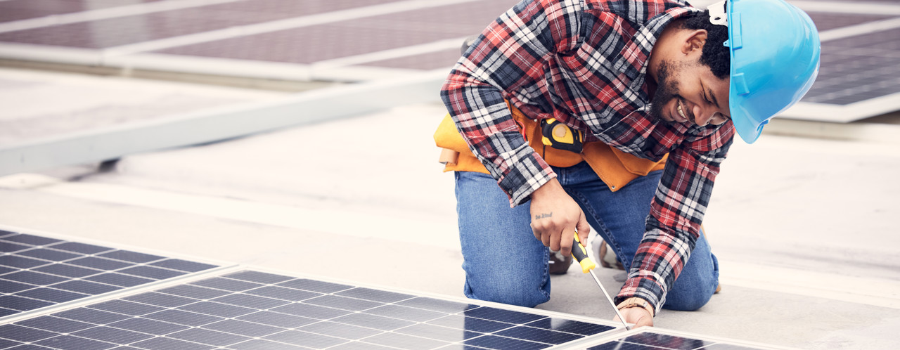 An employee in a blue safety helmet is installing solar panels on a roof, wearing a plaid shirt and a tool belt.