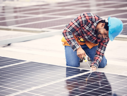 An employee in a blue safety helmet is installing solar panels on a roof, wearing a plaid shirt and a tool belt.