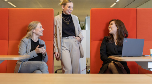 Three women in a modern office setting with orange decor; two are seated at a table while one stands, visibly engaged in conversation.