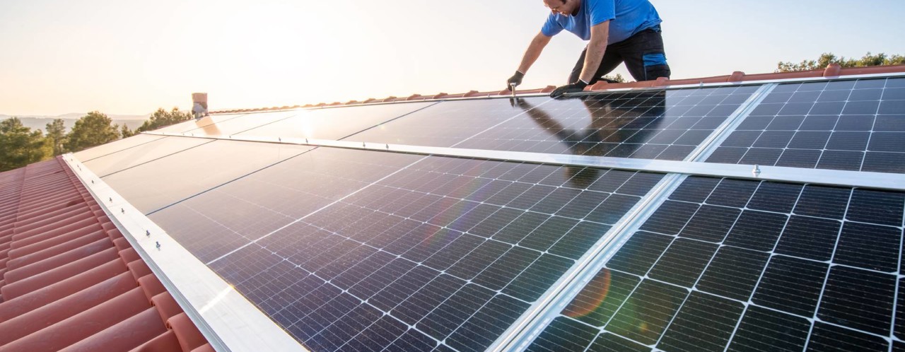 A person installing solar panels on a sloped red tiled roof, with the sun shining in the background.