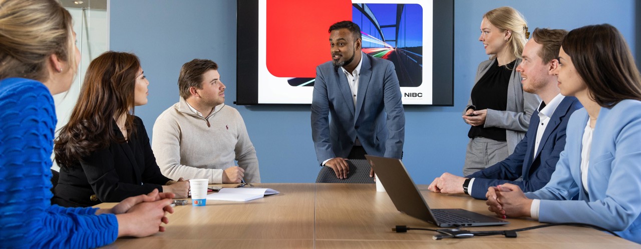 A group of professionals in a meeting room, focused on a presentation displayed on a screen featuring colorful graphic elements.
