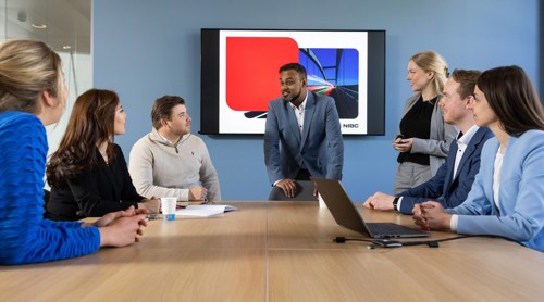 Groep professionals in een vergaderruimte, gefocust op een presentatie op een scherm met kleurrijke grafische elementen.