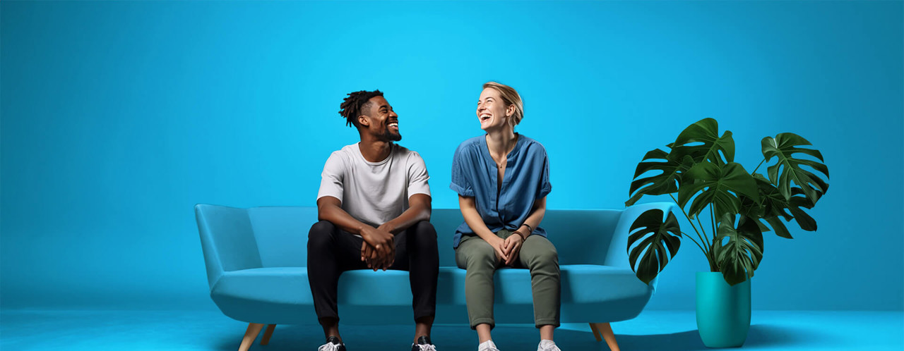 Two people are sitting on a light blue couch against a bright blue background, with a green plant on the floor to the right.