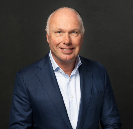 Nick in a dark blue suit with a light blue shirt stands against a black background.