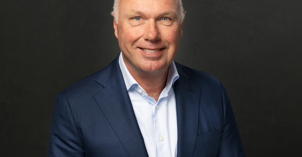 Nick in een donkerblauw pak met een lichtblauw overhemd staat voor een zwarte achtergrond.