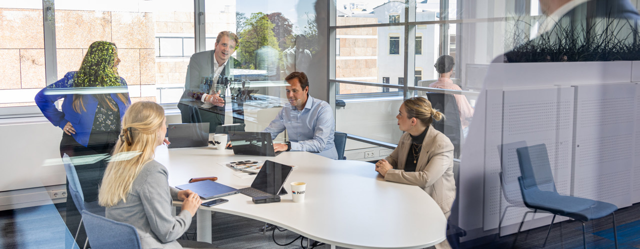 A group of professionals in a meeting room, focused on a presentation with laptops and documents on the table.