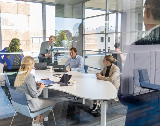 A group of professionals in a meeting room, focused on a presentation with laptops and documents on the table.