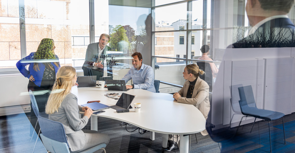 A group of professionals in a meeting room, focused on a presentation with laptops and documents on the table.
