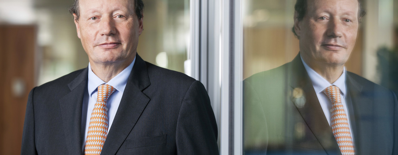 Mr D.M. Sluimers in a black suit with an orange striped tie, standing in front of a glass wall with reflections.