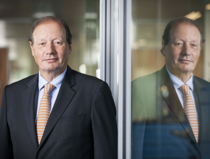 Mr D.M. Sluimers in zwart pak met oranje gestreepte das, staand voor een glazen wand met reflecties.