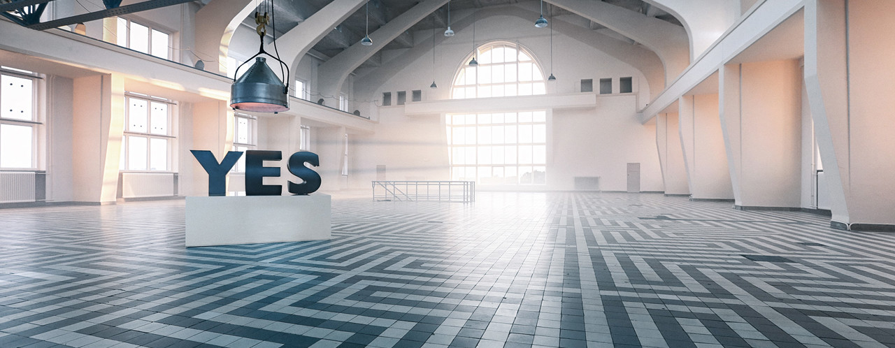 Grote sculptuur met het woord 'JA' in een lege, moderne ruimte met een lichte vloer en veel natuurlijk licht.