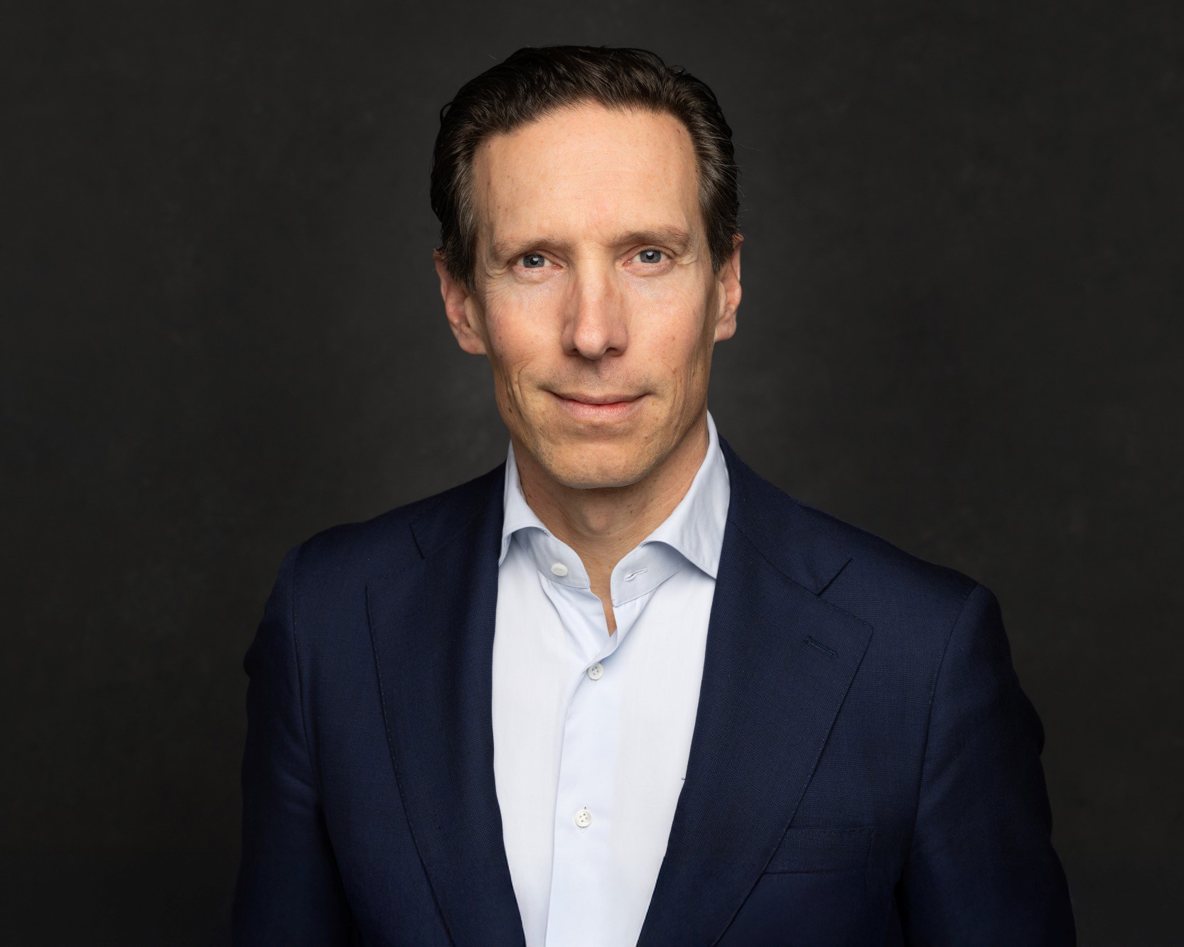 Sven in een donkerblauwe blazer tegen een zwart-grijze achtergrond, met een neutrale blik.