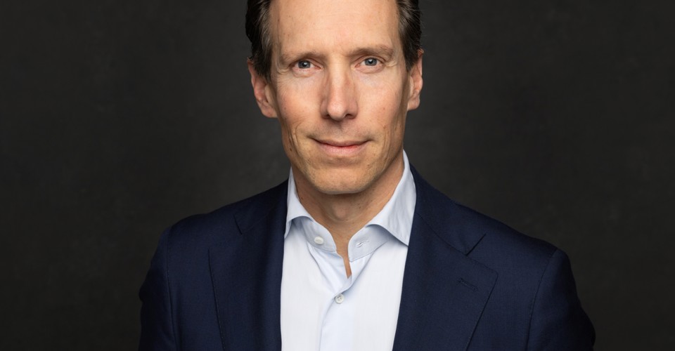 Sven in een donkerblauwe blazer tegen een zwart-grijze achtergrond, met een neutrale blik.