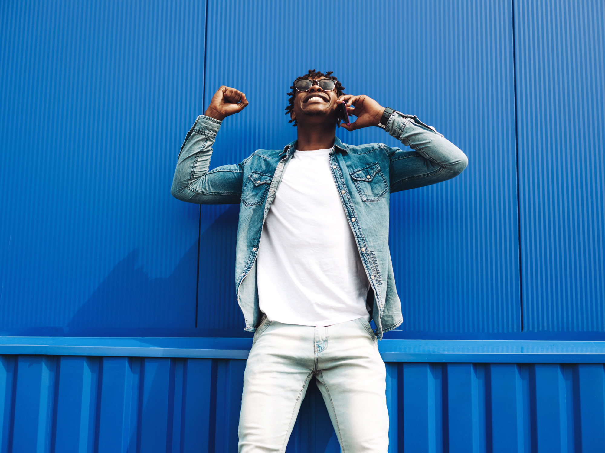 Man in a denim jacket and white T-shirt talking on a mobile phone against a blue wall.