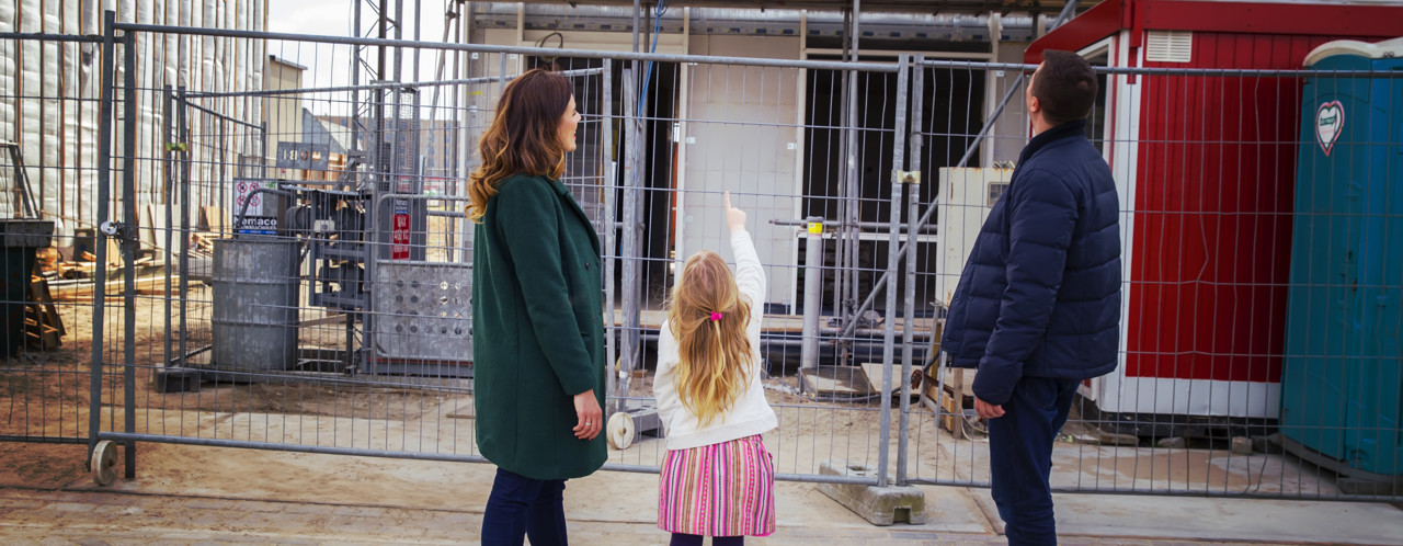 A family is watching a construction site surrounded by a fence; the child is excitedly pointing at something on the site.