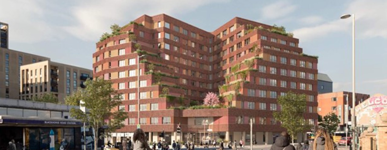 Modern apartment buildings with red facades, surrounded by green plants, with cyclists and pedestrians on the street.