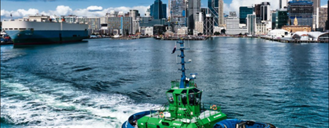 A green tugboat navigates through the water with a modern city skyline in the background, featuring skyscrapers and ships.